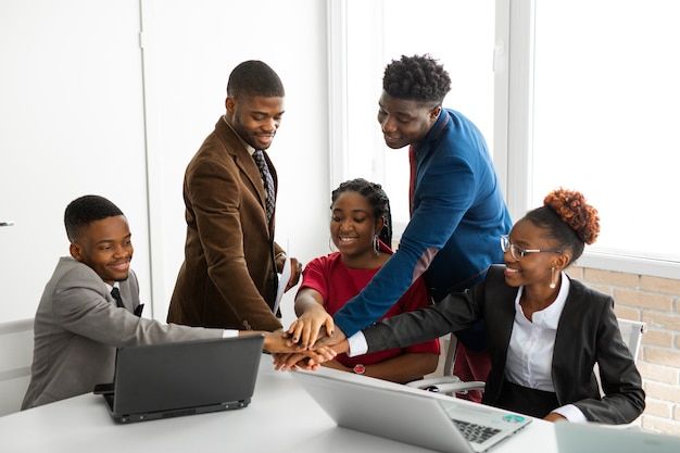 Cinq jeunes professionnels de creatix souriants sont réunis autour d'une table avec des ordinateurs portables. Ils mettent leurs mains ensemble au centre en signe de collaboration et d'esprit d'équipe dans un bureau lumineux.